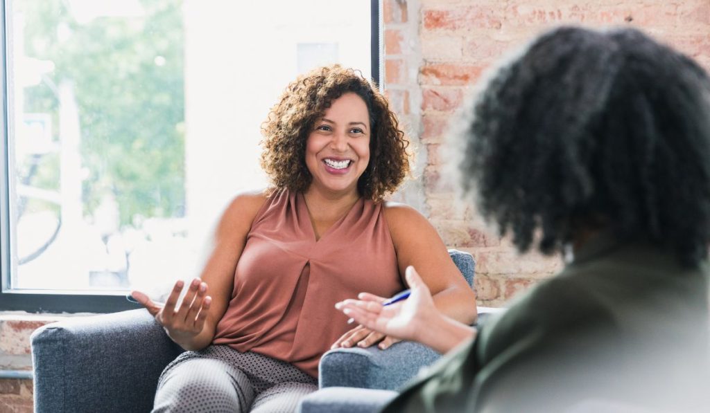 Two women sit in armchairs facing each other, engaged in conversation about different types of therapy for trauma. One woman is smiling and gesturing with her hands while the other has her back to the camera.