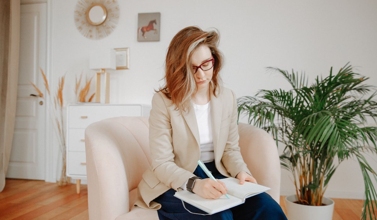 In a room adorned with plants and minimalistic furniture, a person with glasses relaxes in a beige chair. As they write in their notebook, they're perhaps exploring forms of trauma therapy to understand what type of therapy is best for trauma healing.