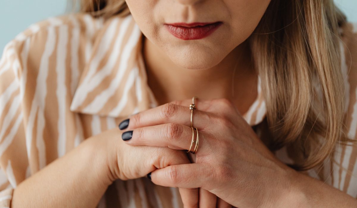 A hypervigilant woman with folded hands wears gold rings and a striped shirt, showing dark nail polish and light brown hair.